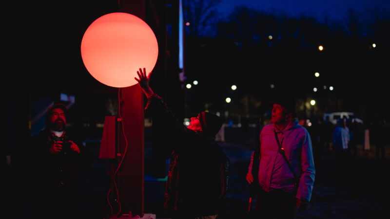 A photograph of an orange glowing orb that is suspended to float in the air outdoors at night. Several people can be seen in the glow and one reaches up to touch it.