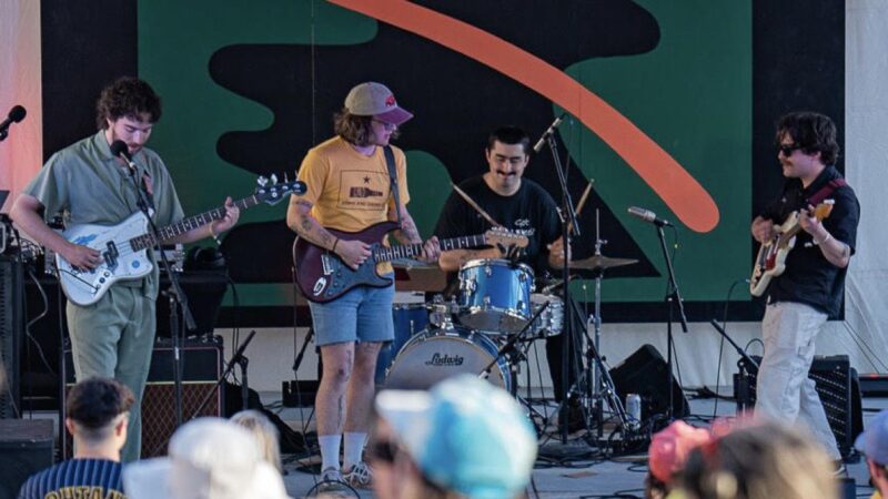 A band consisting of three light skinned men playing guitar and bass and one man playing drums stands on a stage with an abstract green and orange mural in the background