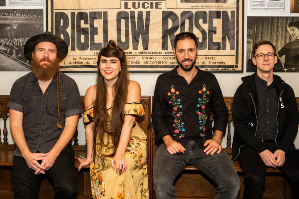 a photograph of a woman, Kat Wright, and three men, members of her band, sitting on a long bench with posters behind them