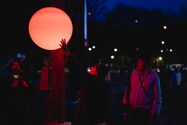 A photograph of an orange glowing orb that is suspended to float in the air outdoors at night. Several people can be seen in the glow and one reaches up to touch it.