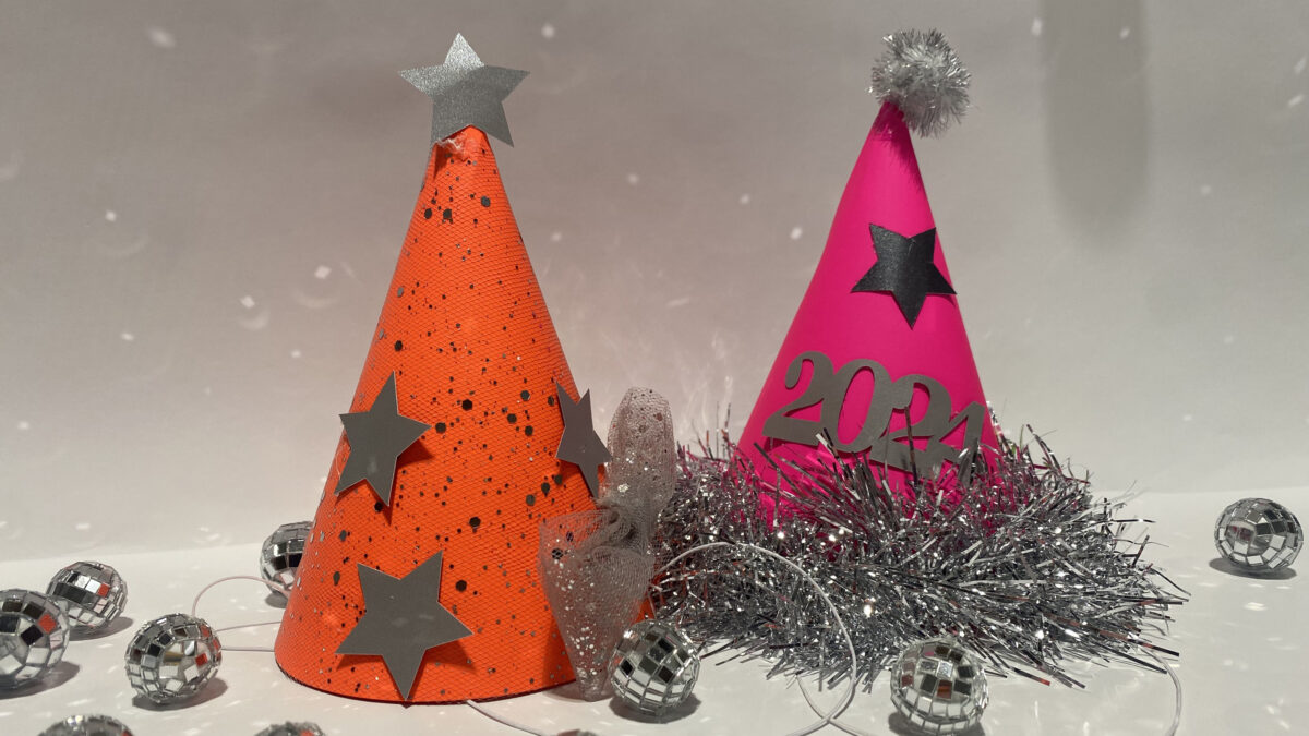 a photo of two homemade conical party hats, one bright orange, one bright pink, both adorned with silver stars, tinsel and glitter