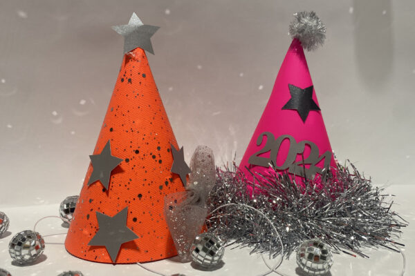 a photo of two homemade conical party hats, one bright orange, one bright pink, both adorned with silver stars, tinsel and glitter