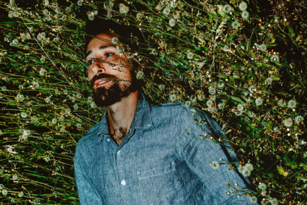 a photograph of Matt Lorenz, aka The Suitcase Junket, a light skinned man with dark hair and a beard, lying on the ground surrounded by dandelions that have gone to seed