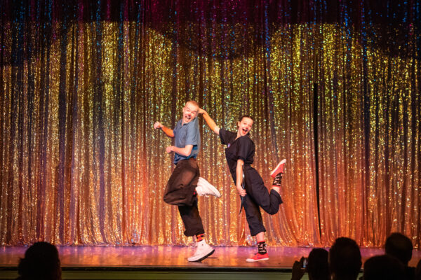 a photograph of Circus Smirkus performing at Highlight New Year's Eve. A light skinned young ma and woman dance in unison on a stage with a sparkly copper colored curtain behind them