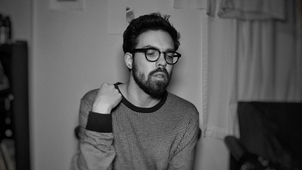 A black and white photograph of a light skinned man with dark hair, a short beard and glasses seen from the chest up
