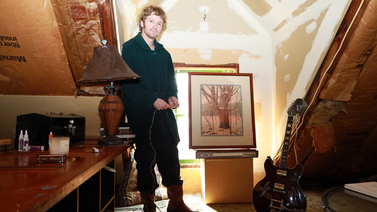Color photo of white man looking at camera in room under construction. Guitar on right, desk with lamp on left, framed photograph in center next to man. slanted ceiling and window behind photograph, white paint on walls, bright light in window brightening room.