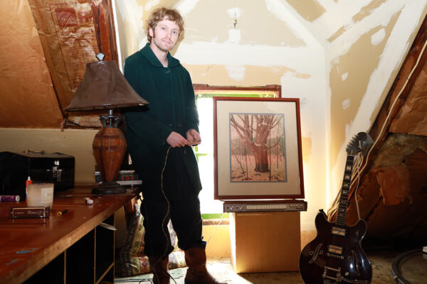Color photo of white man looking at camera in room under construction. Guitar on right, desk with lamp on left, framed photograph in center next to man. slanted ceiling and window behind photograph, white paint on walls, bright light in window brightening room.