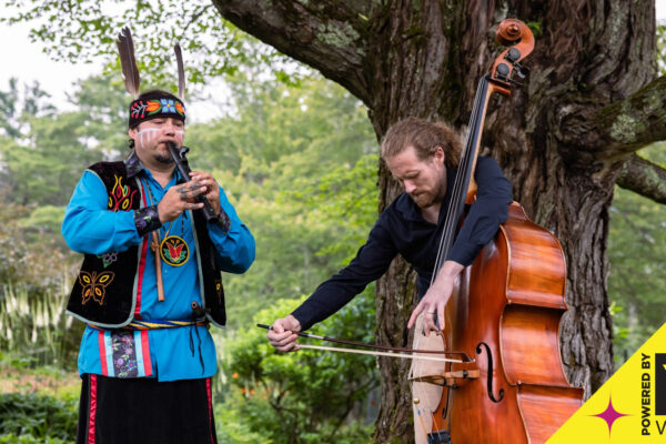 Color photo of two men, one white, one indigenous with medium brown skin dressed in cultural clothing, playing instruments. Background has large tree in front of forest. Text saying "powered by vsecu" in bottom right corner.