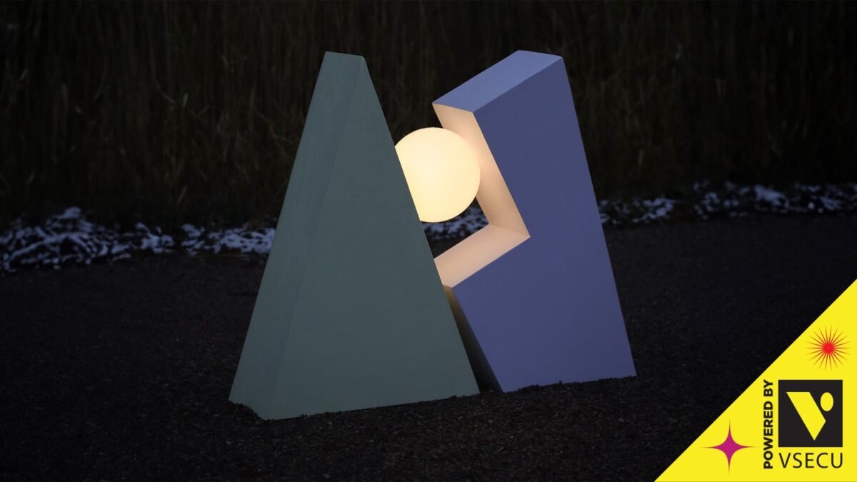 A photograph of a large abstract outdoor sculpture taken at dusk. The sculpture balances a glowing orb between a pale green triangle and another geometric shape
