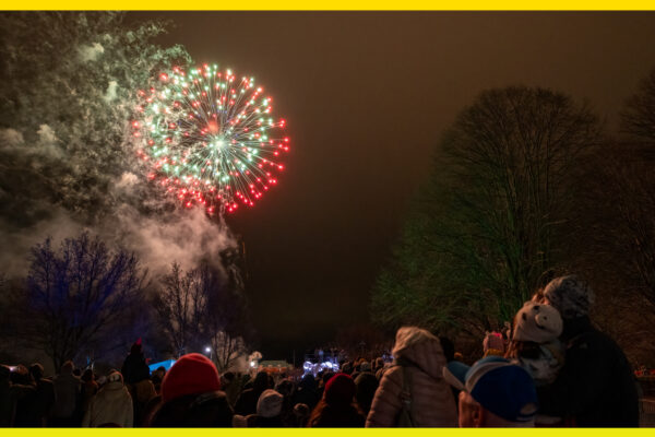 A dark sky fills the image with two colorful explosions in the left corner. In the bottom right corner, there are a few people.