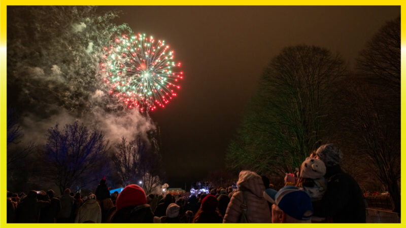 A dark sky fills the image with two colorful explosions in the left corner. In the bottom right corner, there are a few people.