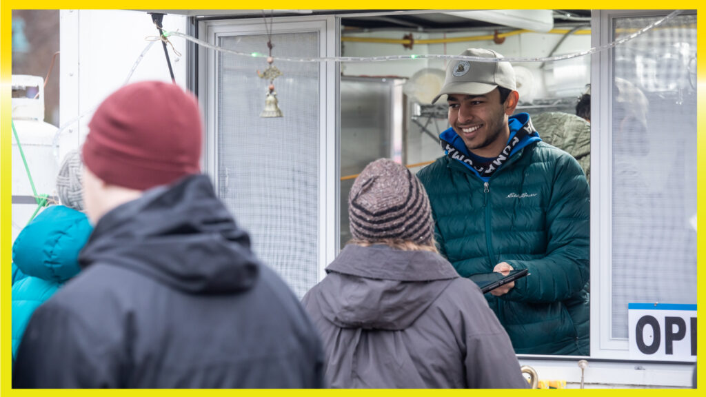 A person with a hat on and coat is serving food out of a sliding window. Two people who are bundled are waiting in line.