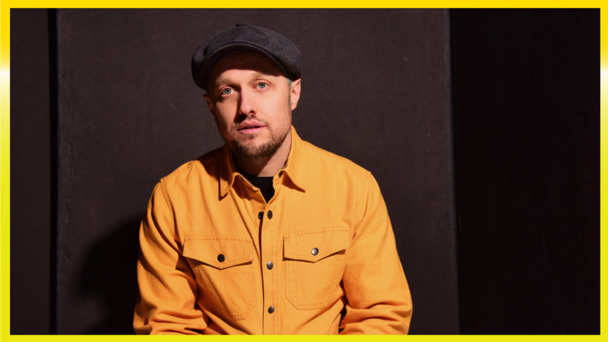 A person with a yellow shirt on, a beard, and a grey hat on with a dark background.