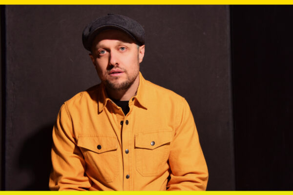 A person with a yellow shirt on, a beard, and a grey hat on with a dark background.