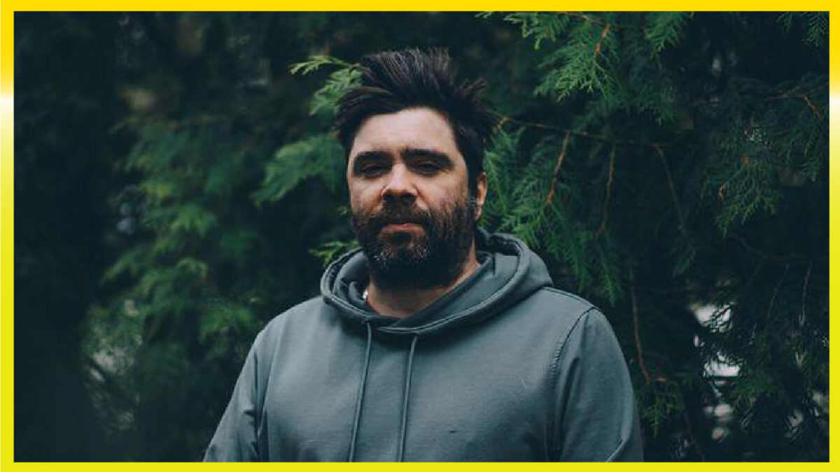 A person who has dark hair and a beard with a grey sweatshirt on. Standing in front of pine trees.