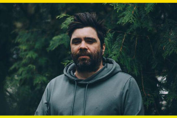 A person who has dark hair and a beard with a grey sweatshirt on. Standing in front of pine trees.