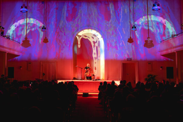 Photo of a cellist performing inside a church with imagery and color being projected behind them.