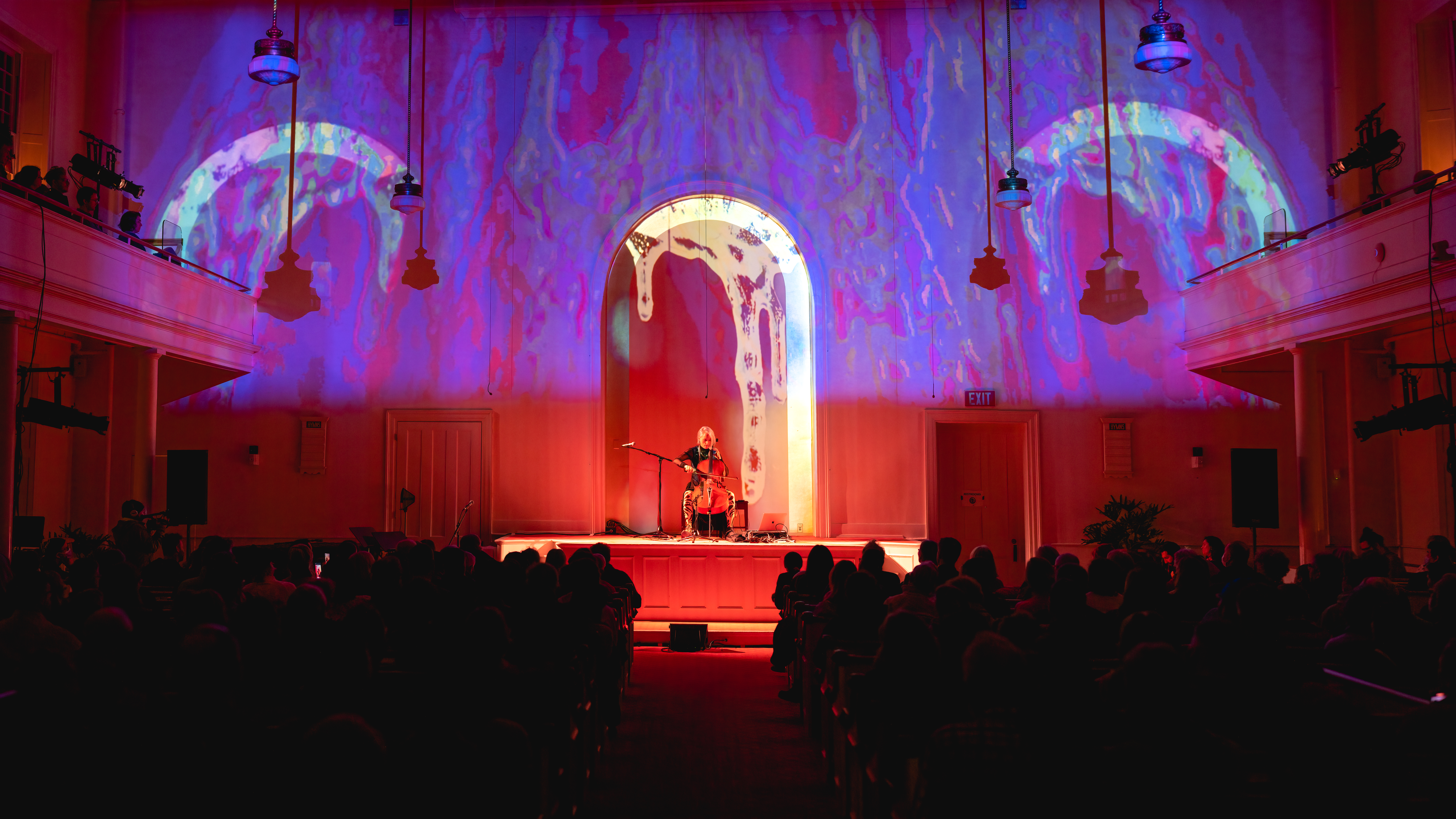 Photo of a cellist performing inside a church with imagery and color being projected behind them.